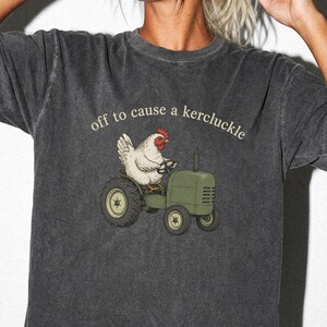 May include: A charcoal gray short-sleeve t-shirt with a graphic print of a white chicken driving a green tractor. Above the chicken, the text reads "off to cause a kercluckle" in a light-colored, curved font. The fabric has a washed-out texture.