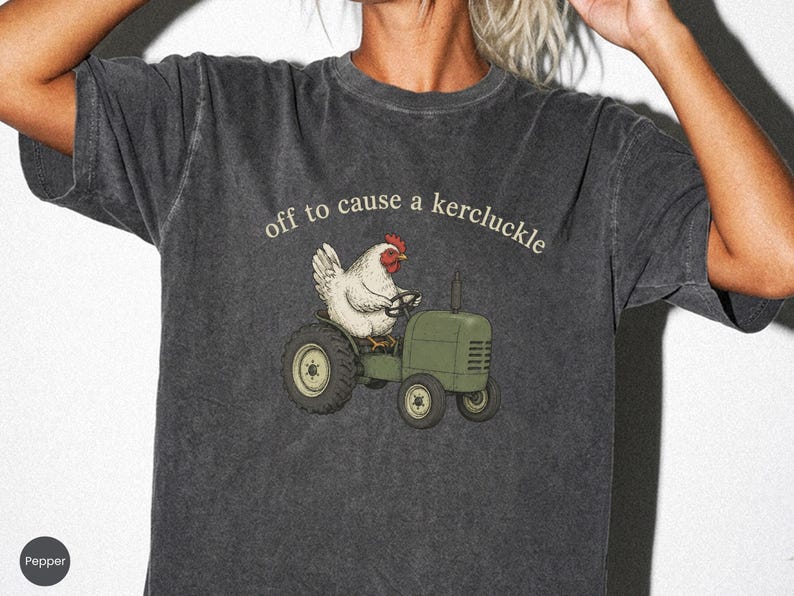 May include: A charcoal gray short-sleeve t-shirt with a graphic print of a white chicken driving a green tractor. Above the chicken, the text reads "off to cause a kercluckle" in a light-colored, curved font. The fabric has a washed-out texture.