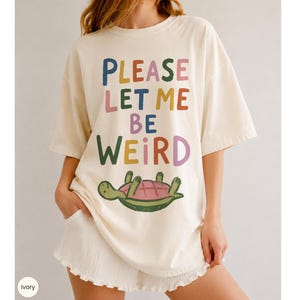 Könnte beinhalten: Ein elfenbeinfarbenes Kurzarm-T-Shirt mit dem mehrfarbigen Text "PLEASE LET ME BE WEIRD" auf der Vorderseite. Unter dem Text befindet sich eine grafische Illustration einer grünen Schildkröte, die auf dem Rücken liegt und einen rosa Panzer hat.