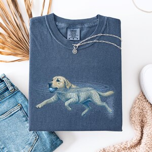 May include: A blue short-sleeved t-shirt featuring a graphic of a light-colored dog swimming in water while holding a blue ball in its mouth. The shirt has a crew neckline and a Comfort Colors brand label printed on the inside of the collar.