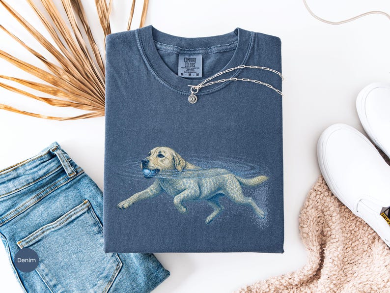 May include: A blue short-sleeved t-shirt featuring a graphic of a light-colored dog swimming in water while holding a blue ball in its mouth. The shirt has a crew neckline and a Comfort Colors brand label printed on the inside of the collar.