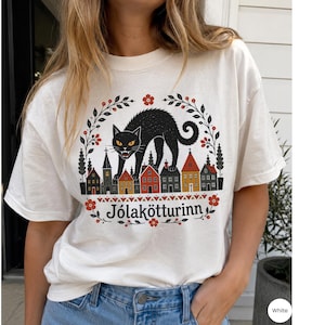 May include: A white short-sleeve t-shirt featuring a graphic of a black cat standing over a row of colorful houses. The design is framed by floral branches and includes the text J&oacute;lak&ouml;tturinn in black lettering centered below the houses.