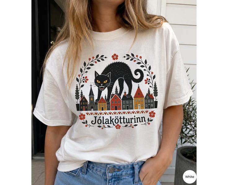 May include: A white short-sleeve t-shirt featuring a graphic of a black cat standing over a row of colorful houses. The design is framed by floral branches and includes the text J&oacute;lak&ouml;tturinn in black lettering centered below the houses.