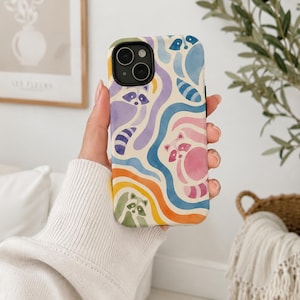 Puede incluir: Una funda para smartphone con un patr&oacute;n estilo acuarela de mapaches integrados en bandas onduladas y fluidas de colores azul, morado, rosa, naranja y verde. El dise&ntilde;o cubre la parte posterior de la funda, que tiene un recorte negro para la c&aacute;mara.