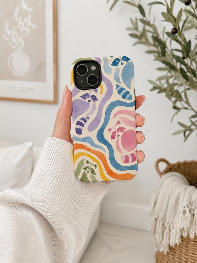 Puede incluir: Una funda para smartphone con un patr&oacute;n estilo acuarela de mapaches integrados en bandas onduladas y fluidas de colores azul, morado, rosa, naranja y verde. El dise&ntilde;o cubre la parte posterior de la funda, que tiene un recorte negro para la c&aacute;mara.