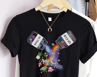 Peut inclure: T-shirt noir avec un graphique de deux bocaux étiquetés "Autisme" et "Queer" qui versent des paillettes et des étoiles colorées. Le graphique comprend également un arc-en-ciel, un chat, un champignon, une grenouille et une pieuvre.