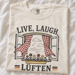 Pode incluir: Uma t-shirt cor de marfim com um gráfico de uma janela aberta com cortinas aos quadrados vermelhos e brancos, uma floreira com flores coloridas e o texto "LIVE, LAUGH, LÜFTEN" com pequenas bandeiras alemãs em ambos os lados da palavra LÜFTEN.