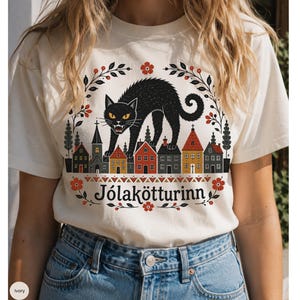 May include: An ivory t-shirt featuring a graphic illustration of a black cat arching its back over a row of small houses. The design includes red flowers, dark green foliage, and the text J&oacute;lak&ouml;tturinn printed in a black serif font.