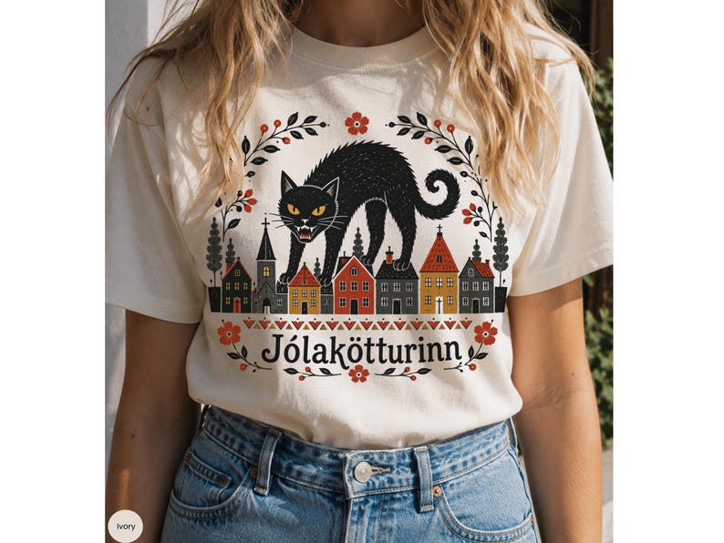 May include: An ivory t-shirt featuring a graphic illustration of a black cat arching its back over a row of small houses. The design includes red flowers, dark green foliage, and the text J&oacute;lak&ouml;tturinn printed in a black serif font.