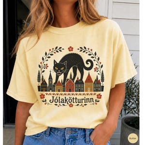May include: A pale yellow t-shirt featuring a graphic print of a black cat arching its back over a row of small houses. The design is framed by a floral wreath and includes the text J&oacute;lak&ouml;tturinn in a black serif font at the bottom center.