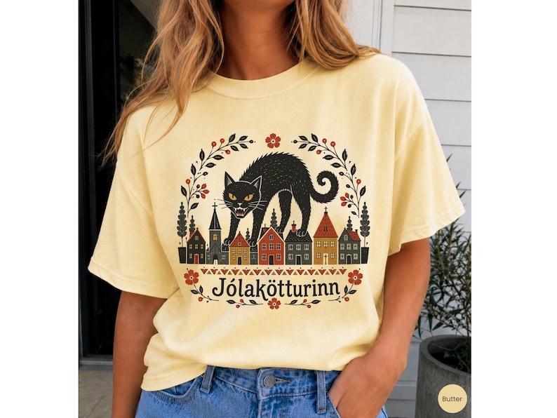 May include: A pale yellow t-shirt featuring a graphic print of a black cat arching its back over a row of small houses. The design is framed by a floral wreath and includes the text J&oacute;lak&ouml;tturinn in a black serif font at the bottom center.