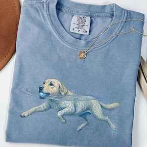 May include: A blue jean colored t-shirt with a graphic of a light-colored dog running with a blue ball in its mouth. The shirt features a Comfort Colors brand label at the neckline, which indicates a size small and 100% cotton material composition.