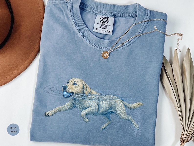 May include: A blue jean colored t-shirt with a graphic of a light-colored dog running with a blue ball in its mouth. The shirt features a Comfort Colors brand label at the neckline, which indicates a size small and 100% cotton material composition.