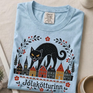May include: A light blue t-shirt featuring a graphic of a large black cat arching its back over a row of colorful houses. The design includes floral accents and the text J&oacute;lak&ouml;tturinn. The shirt has a Comfort Colors brand label at the neck.
