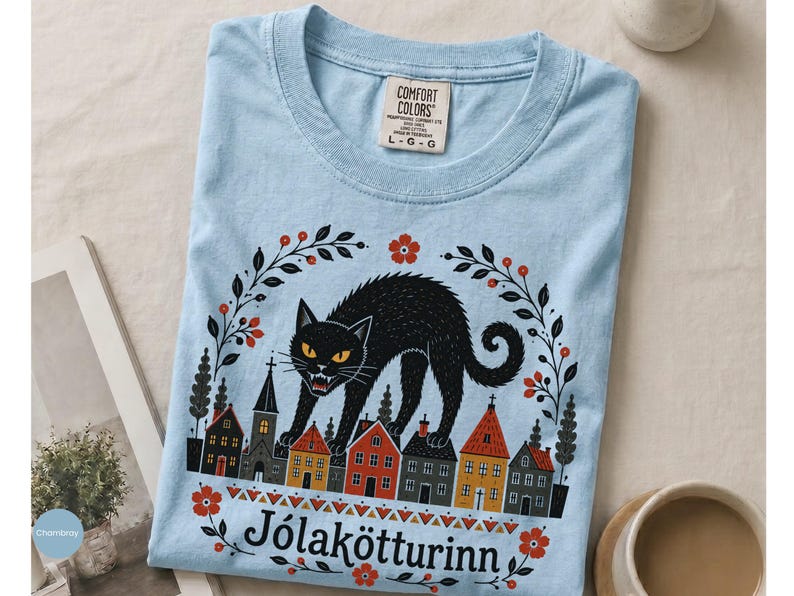 May include: A light blue t-shirt featuring a graphic of a large black cat arching its back over a row of colorful houses. The design includes floral accents and the text J&oacute;lak&ouml;tturinn. The shirt has a Comfort Colors brand label at the neck.
