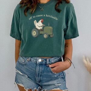 May include: A dark teal short-sleeve t-shirt featuring a graphic of a white chicken driving a green tractor. The text above the chicken reads, "off to cause a kercluckle" in a simple white font. The shirt has a crew neckline and a relaxed fit.