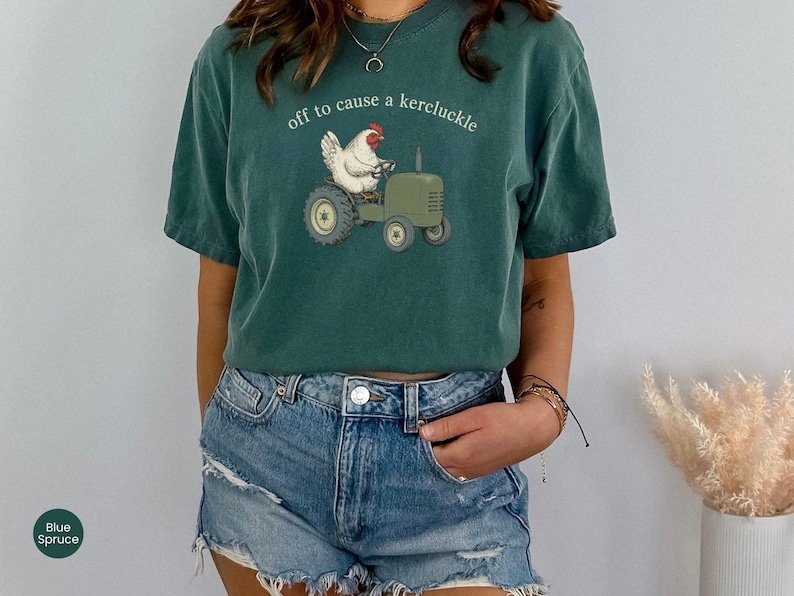 May include: A dark teal short-sleeve t-shirt featuring a graphic of a white chicken driving a green tractor. The text above the chicken reads, "off to cause a kercluckle" in a simple white font. The shirt has a crew neckline and a relaxed fit.