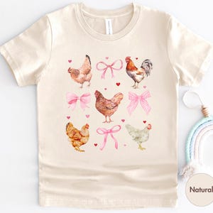 May include: A beige t-shirt with a graphic print of chickens wearing pink bows. The shirt has a crew neck and short sleeves.