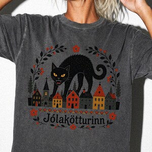 May include: A charcoal gray short-sleeve t-shirt featuring a graphic of a black cat standing over a row of small houses. The design includes red flowers, dark leaves, and the text "J&oacute;lak&ouml;tturinn" printed in black below the houses.