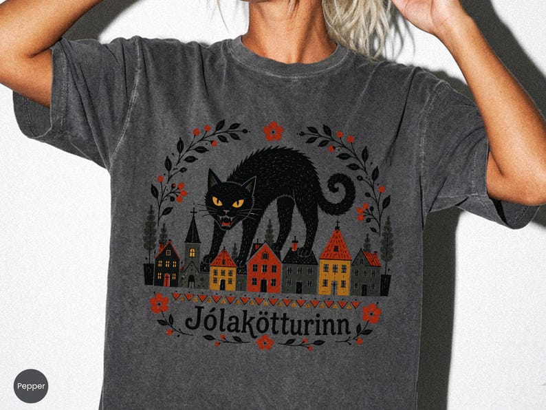 May include: A charcoal gray short-sleeve t-shirt featuring a graphic of a black cat standing over a row of small houses. The design includes red flowers, dark leaves, and the text "J&oacute;lak&ouml;tturinn" printed in black below the houses.