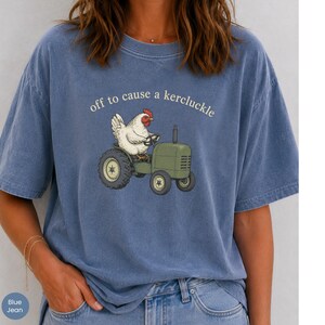 May include: A blue short-sleeve t-shirt featuring a graphic of a white chicken driving a green tractor. Above the chicken, the text reads "off to cause a kercluckle" in a simple white font. The shirt has a crew neckline and a relaxed fit.