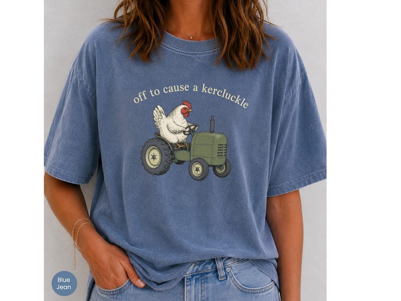 May include: A blue short-sleeve t-shirt featuring a graphic of a white chicken driving a green tractor. Above the chicken, the text reads "off to cause a kercluckle" in a simple white font. The shirt has a crew neckline and a relaxed fit.