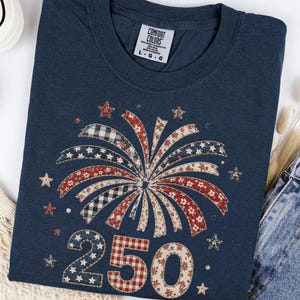 May include: A navy blue t-shirt featuring a graphic of a firework made of patterned ribbons in red, white, and blue, with the number 250 below it in a similar patchwork style. The shirt has a Comfort Colors label showing size L.