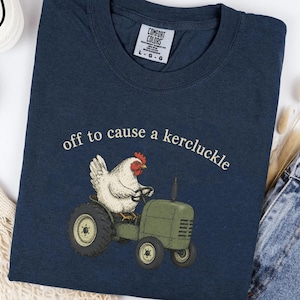 May include: A navy blue crew neck t-shirt featuring a graphic of a white chicken driving a green tractor. Above the chicken, the text reads "off to cause a kercluckle" in a light-colored font. The shirt has a Comfort Colors brand label.