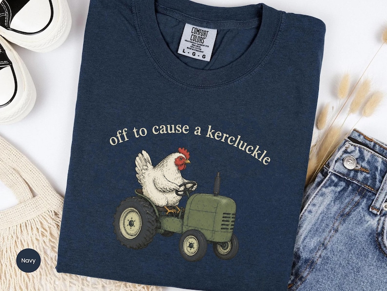 May include: A navy blue crew neck t-shirt featuring a graphic of a white chicken driving a green tractor. Above the chicken, the text reads "off to cause a kercluckle" in a light-colored font. The shirt has a Comfort Colors brand label.