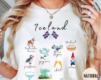 May include: White t-shirt with a graphic design of various Icelandic icons, including horses, geysers, puffins, the Blue Lagoon, and the Northern Lights. The text "Iceland" is at the top of the design, and the text "skál!" is at the bottom.