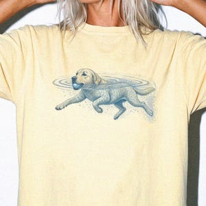 May include: A pale yellow short-sleeve t-shirt featuring a blue graphic illustration of a dog swimming in water while holding a ball in its mouth. The design includes ripples in the water surrounding the dog and small blue speckles below it.
