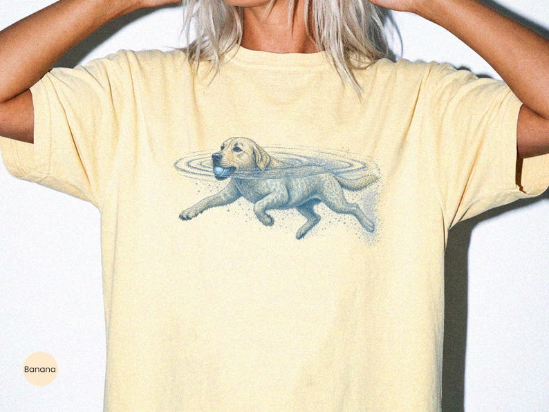 May include: A pale yellow short-sleeve t-shirt featuring a blue graphic illustration of a dog swimming in water while holding a ball in its mouth. The design includes ripples in the water surrounding the dog and small blue speckles below it.