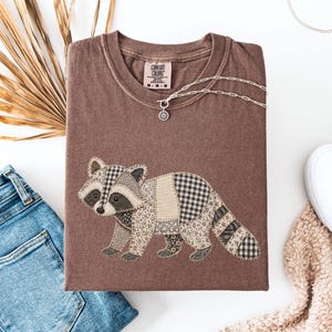 May include: A brown short-sleeved t-shirt featuring a graphic of a raccoon composed of various patterned fabric patches. The shirt has a crew neckline with a Comfort Colors label and is displayed with a silver chain necklace.