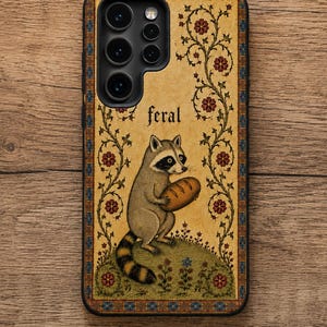 K&ouml;nnte beinhalten: Eine rechteckige Handyh&uuml;lle mit beigem Hintergrund und einer zentralen Illustration eines Waschb&auml;ren, der einen Laib Brot h&auml;lt. Das Wort feral ist &uuml;ber dem Waschb&auml;ren aufgedruckt. Das Design umfasst florale Ranken und eine blau-orange Schmuckbord&uuml;re.