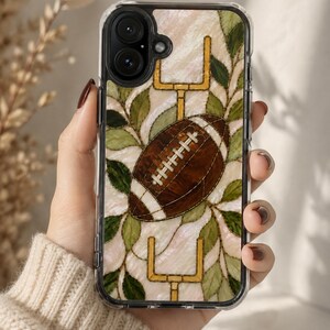 May include: A clear phone case featuring a stained glass style design with a brown football in the center, flanked by yellow goalposts and green leaves against a pearlescent white background. The case has a black camera lens cutout.