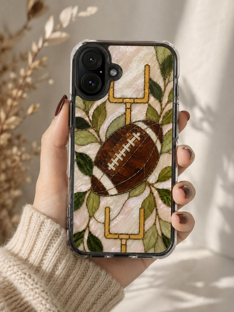 May include: A clear phone case featuring a stained glass style design with a brown football in the center, flanked by yellow goalposts and green leaves against a pearlescent white background. The case has a black camera lens cutout.