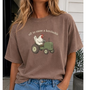 May include: A brown short-sleeve t-shirt featuring a graphic of a white chicken driving a green tractor. Above the chicken, the text reads "off to cause a kercluckle" in a simple white font. The shirt has a crew neckline and a relaxed fit.