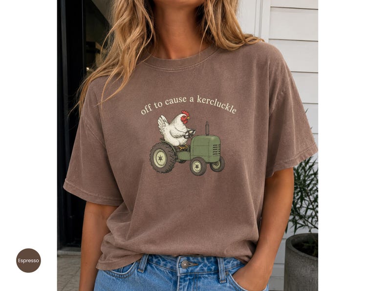 May include: A brown short-sleeve t-shirt featuring a graphic of a white chicken driving a green tractor. Above the chicken, the text reads "off to cause a kercluckle" in a simple white font. The shirt has a crew neckline and a relaxed fit.