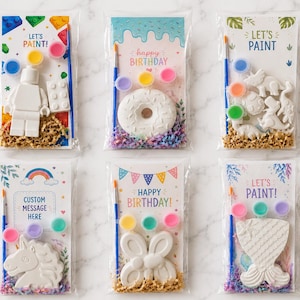 May include: Six clear plastic bags containing paint-your-own plaster figures, including a block figure, donut, dinosaurs, unicorn, butterfly, and mermaid tail. Each kit includes a paintbrush, three small paint pots, and a printed card with themes.