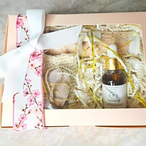 May include: A gift box with a white ribbon and floral accents. Inside, a bottle of serum, heart-shaped stones, and bath products are nestled in decorative paper. The serum bottle has a gold cap and a label that reads "Radiance".