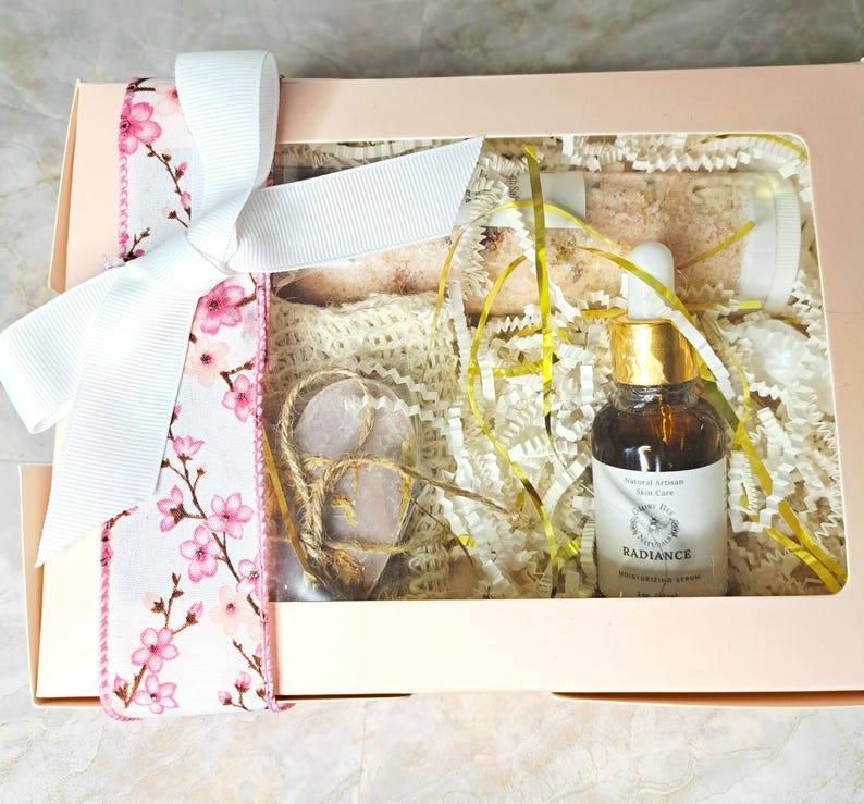 May include: A gift box with a white ribbon and floral accents. Inside, a bottle of serum, heart-shaped stones, and bath products are nestled in decorative paper. The serum bottle has a gold cap and a label that reads "Radiance".