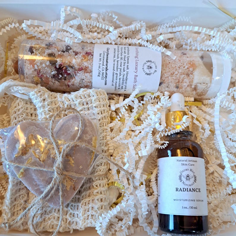 May include: A gift set containing a clear tube of bath salts, a heart-shaped soap, and a moisturizing serum. The bath salts are labeled "Soothing Luxury Bath Salts" and the serum is labeled "Radiance". The set is packaged with white paper filler.