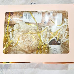 May include: A gift box with a clear window, containing a heart-shaped soap, a glass tube of bath salts, and a small amber bottle. The box is filled with gold and white crinkle paper and gold ribbon. The soap is decorated with dried flowers.