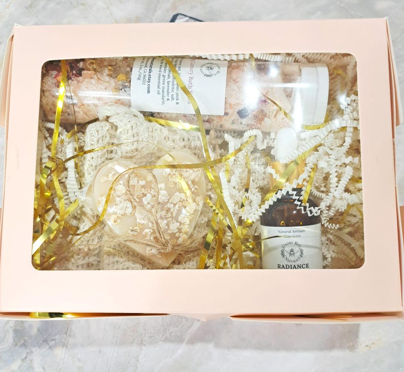 May include: A gift box with a clear window, containing a heart-shaped soap, a glass tube of bath salts, and a small amber bottle. The box is filled with gold and white crinkle paper and gold ribbon. The soap is decorated with dried flowers.