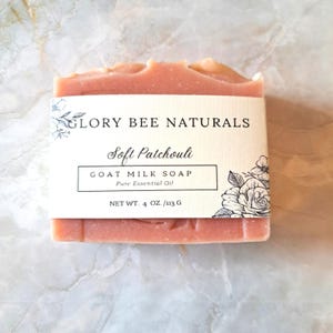 May include: A bar of goat milk soap with a light pink hue. The label reads "GLORY BEE NATURALS Soft Patchouli GOAT MILK SOAP Pure Essential Oil." The net weight is 4 oz. Floral illustrations adorn the label's corners.