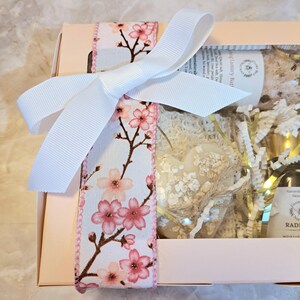 May include: A gift box tied with a white ribbon and a floral ribbon featuring pink cherry blossoms. The box contains a heart-shaped soap, bath salts, and a bottle of moisturizer. The box is light pink.