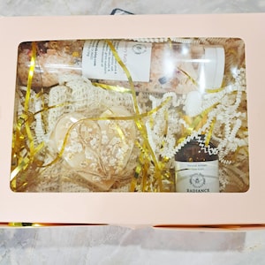 May include: A gift box with a clear window, containing a heart-shaped soap, a bottle of essential oil, and a tube of bath salts. The box is filled with gold and white decorative filler. The essential oil bottle has a label that reads "Radiance".