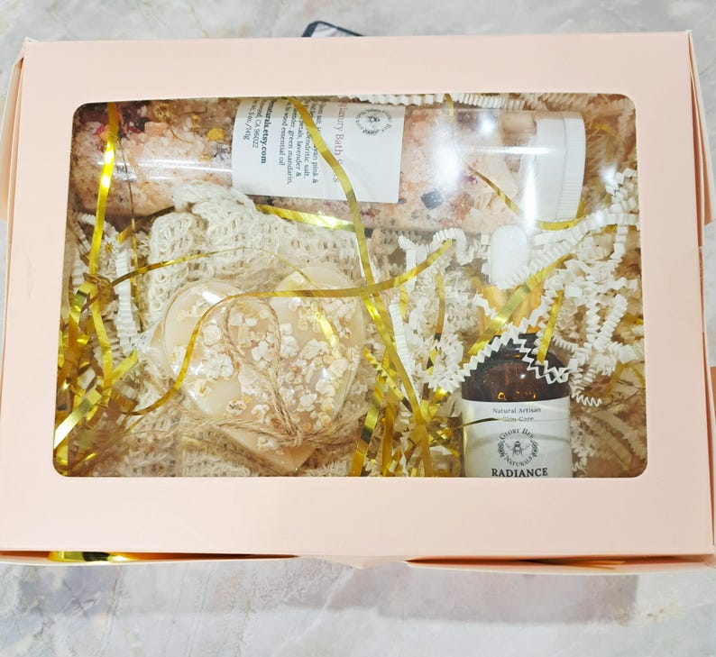 May include: A gift box with a clear window, containing a heart-shaped soap, a bottle of essential oil, and a tube of bath salts. The box is filled with gold and white decorative filler. The essential oil bottle has a label that reads "Radiance".