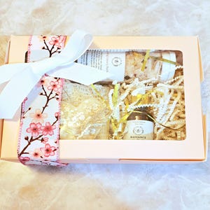 May include: A gift box with a clear window, tied with a white ribbon and a floral cherry blossom ribbon. Inside are bath products, including a heart-shaped soap, a jar of bath salts, and a bottle of Radiance foot care.