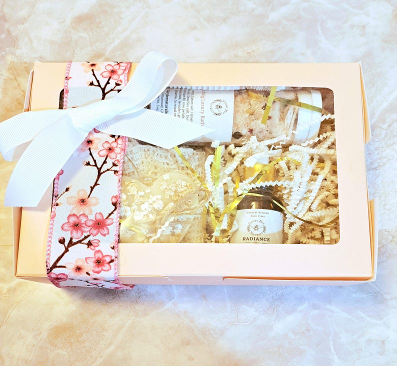 May include: A gift box with a clear window, tied with a white ribbon and a floral cherry blossom ribbon. Inside are bath products, including a heart-shaped soap, a jar of bath salts, and a bottle of Radiance foot care.